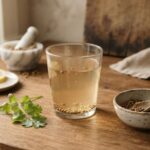 Coriander seed water in a clear glass with floating seeds on a wooden kitchen counter. Prepared by soaking whole coriander seeds overnight. Natural morning drink for blood sugar, cholesterol, and digestion support.